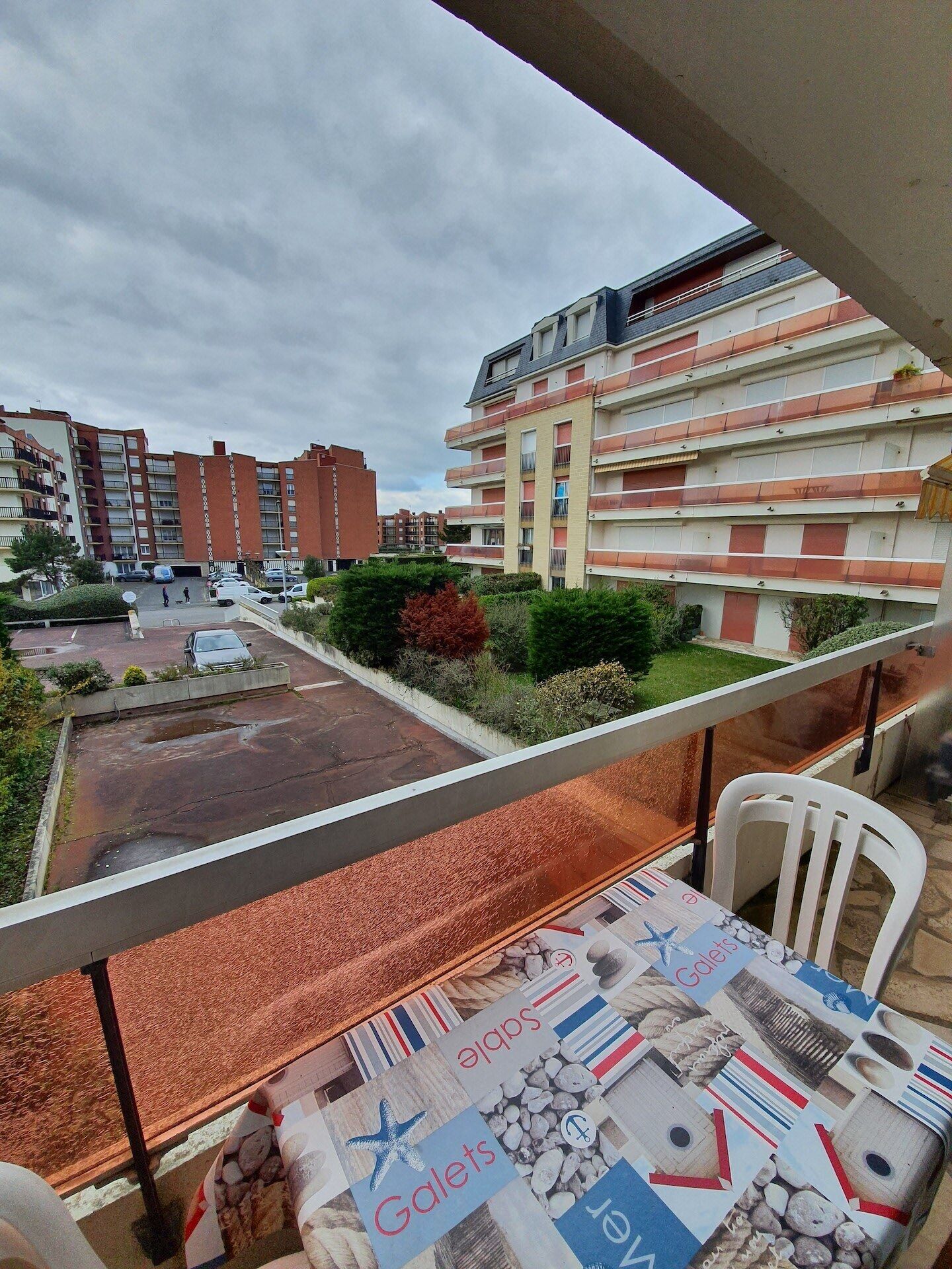 A balcony with a table covered in a nautical-themed tablecloth, overlooking a residential area with multi-story buildings and a parking lot.