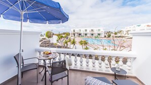 A balcony with a blue umbrella, two chairs, a small table with fruit, and a view of a pool and buildings. - Casa Anemona with terrace, shared pool, sea views and free wifi (Puerto del Carmen)