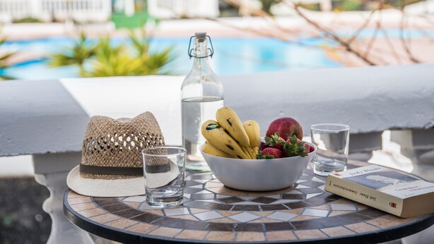 A table with a hat, a bottle, a bowl of fruit, a glass, and a book by J.K. Rowling, set against a poolside view. - Casa Anemona with terrace, shared pool, sea views and free wifi (Puerto del Carmen)