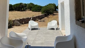 An outdoor patio with a pergola, two white plastic chairs, and a view of a dry landscape with trees.