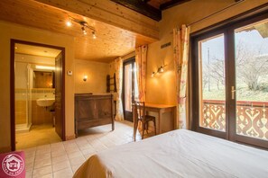 Un dormitorio con cama, un escritorio de madera, una silla, una mesita, un espejo y un baño con lavabo y ducha.