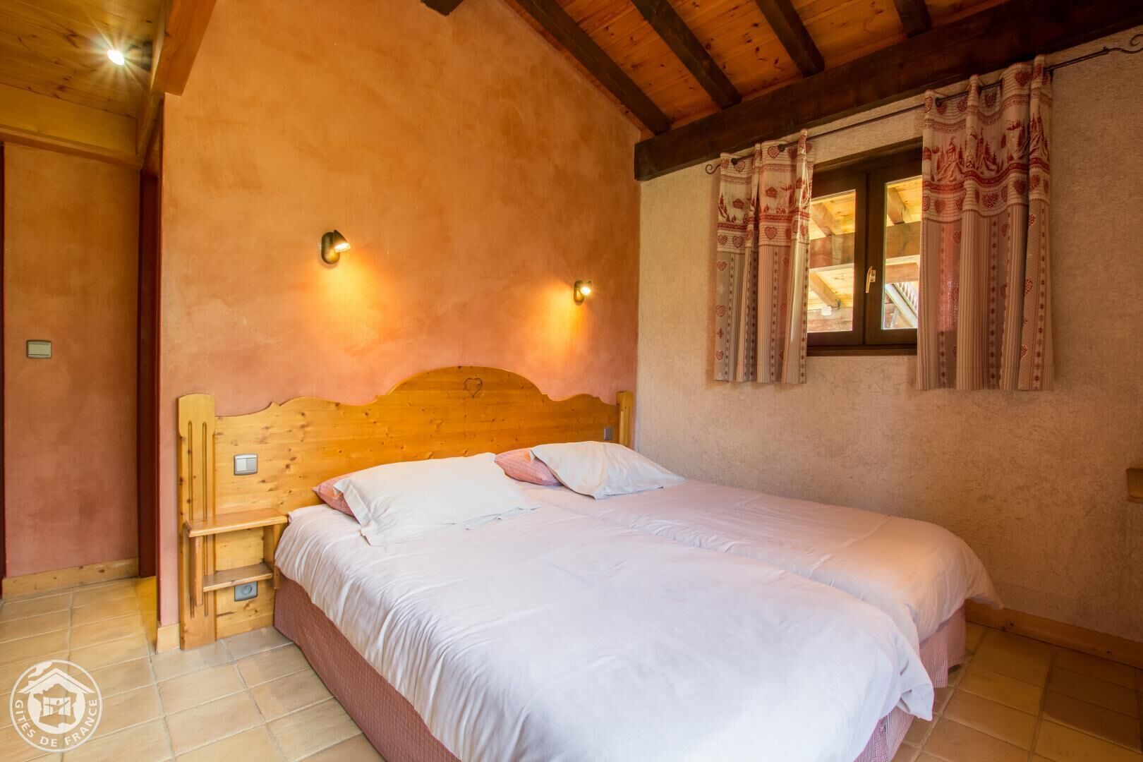 A bedroom with a wooden headboard, a bed with white bedding, two wall-mounted lights, and a window with patterned curtains.