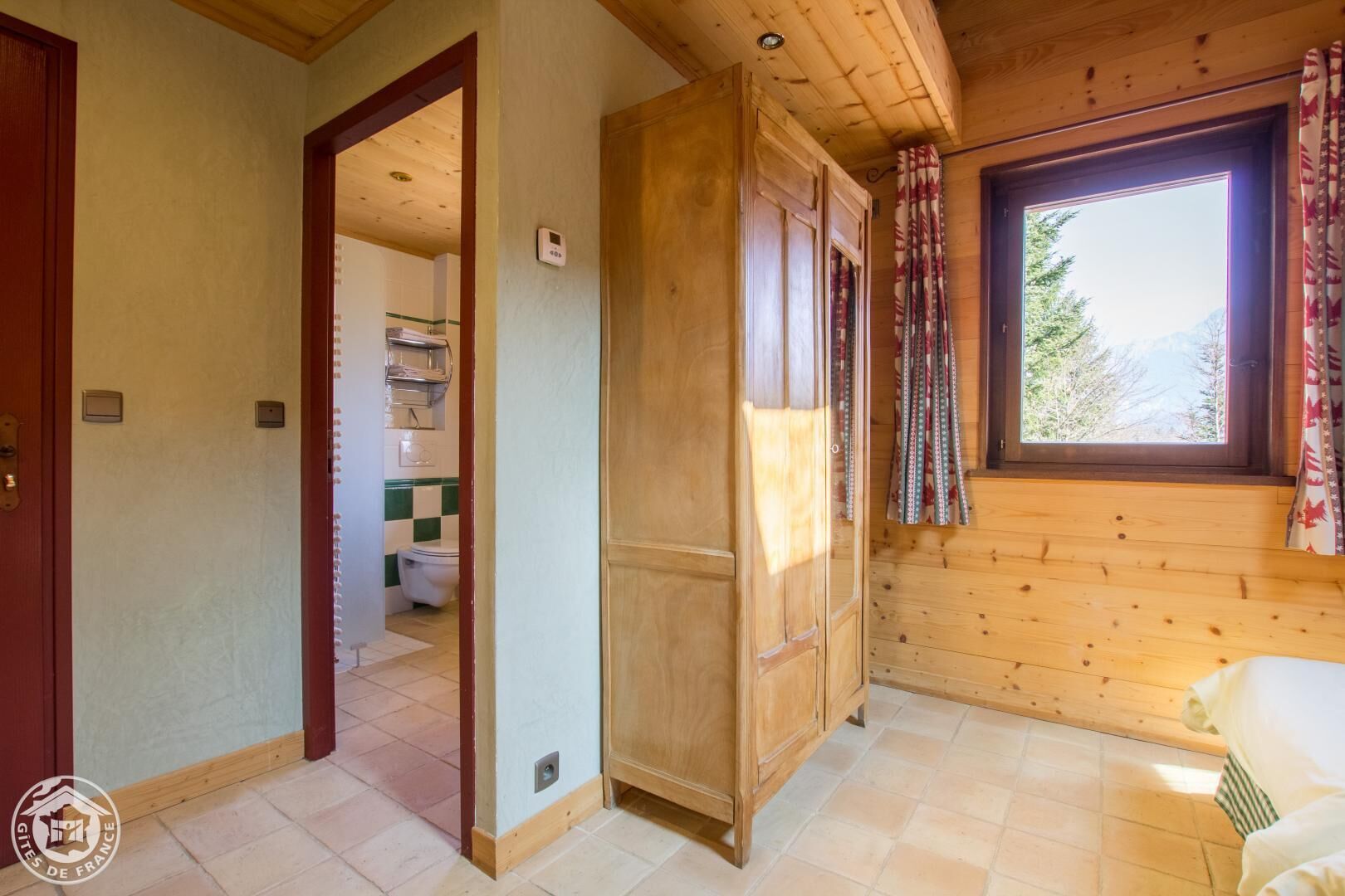 Interior de una cabaña de madera con un baño visible a través de una puerta abierta, una ventana con cortinas y piso de baldosas.