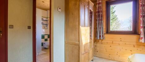 Interior de una cabaña de madera con un baño visible a través de una puerta abierta, una ventana con cortinas y piso de baldosas.