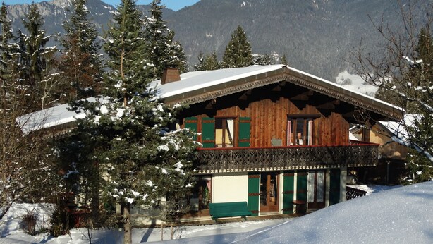 A wooden chalet with a balcony, surrounded by snow-covered trees and mountains in the background.