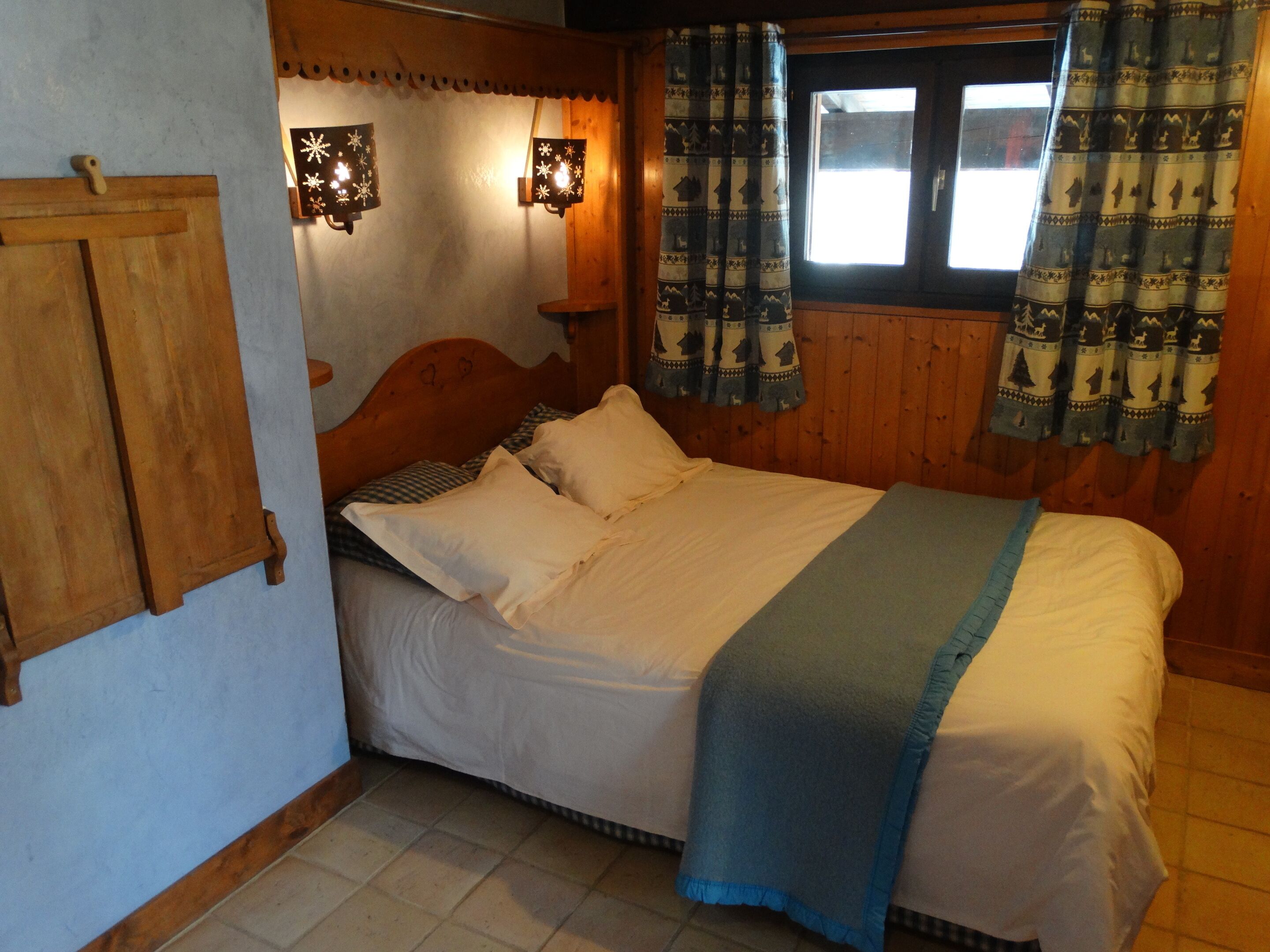 A cozy bedroom with a wooden headboard, a bed with white and blue bedding, two wall-mounted lights, and patterned curtains.