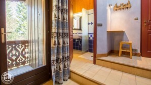 A hotel room with a sliding glass door, a wooden bench, a small stool, a bathroom with a sink and mirror, and a wall-mounted decoration.