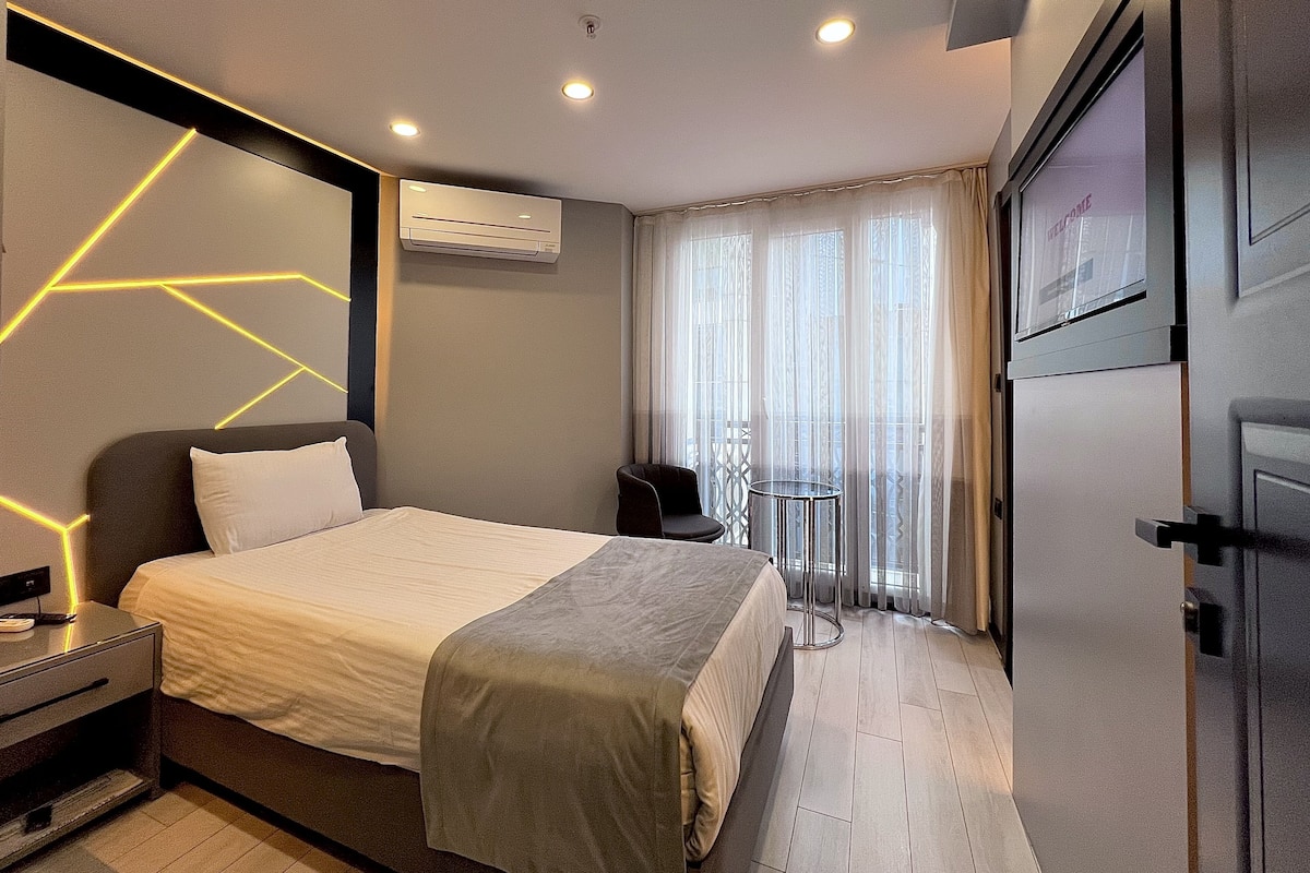 A modern hotel room with a large bed, bedside tables, an air conditioning unit, a small table with a chair, and a view through sheer curtains.