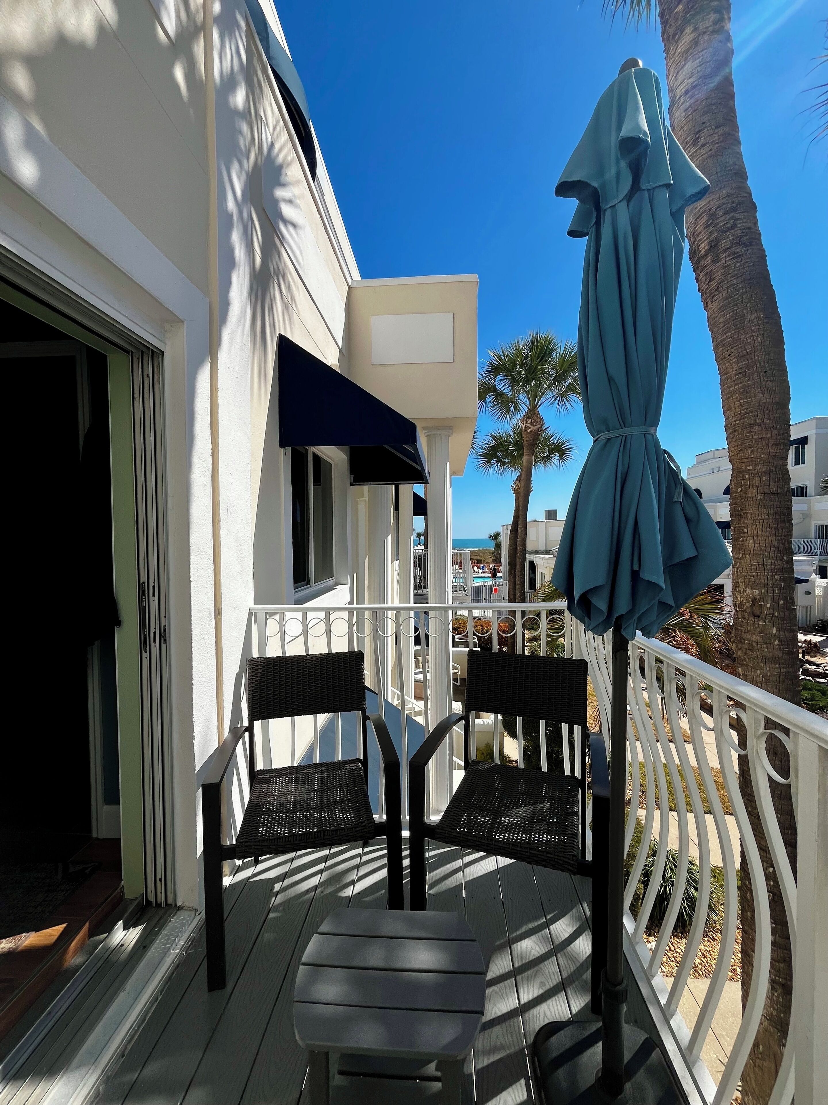A balcony with two wicker chairs and a table, a blue umbrella, and a white railing.