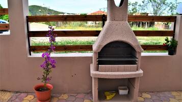 Una parrilla de piedra al aire libre con una ventana al fondo, una maceta con una flor púrpura y un patio con baldosas.