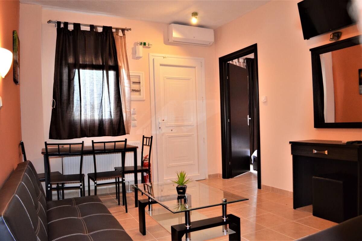A living room with a sofa, a glass-top coffee table, a dining table with chairs, a TV mounted on the wall, and a door leading to another room.