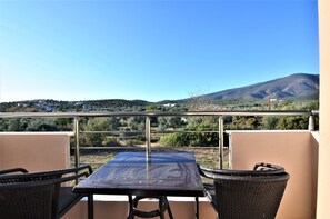Un balcón con mesa y sillas, con vistas a un paisaje montañoso de vegetación escasa y cielo azul despejado.