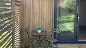 Una terraza de madera con una mesa, sillas, una planta en maceta y un ventanal corredero que da a una zona verde.