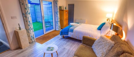 A bedroom with a bed, a sofa, a small table, a wooden cabinet, and a sliding glass door leading to a balcony.