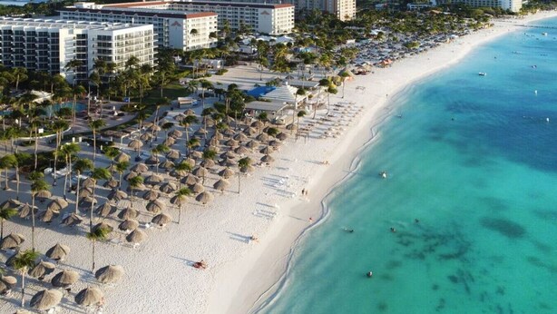 Ein Strandresort mit mehreren Hotelgebäuden, einem Sandstrand gesäumt von Palmen und Strohdächern sowie klarem, türkisfarbenem Wasser.