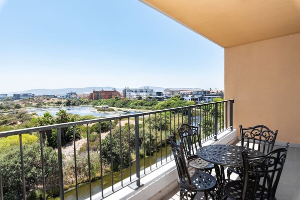 A balcony with a table and chairs, overlooking a river and greenery with buildings in the background. - Modern and Light Apartment in Century City (Cape Town)