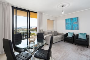 Ein modernes Wohnzimmer mit Glastisch, schwarzen Stühlen, einem grauen Sofa und einem Balkon mit Blick auf die Stadt.