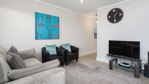 A living room with a beige sofa, two black armchairs, a blue abstract painting, a black clock, and a flat-screen TV on a glass stand. - Modern and Light Apartment in Century City (Cape Town)