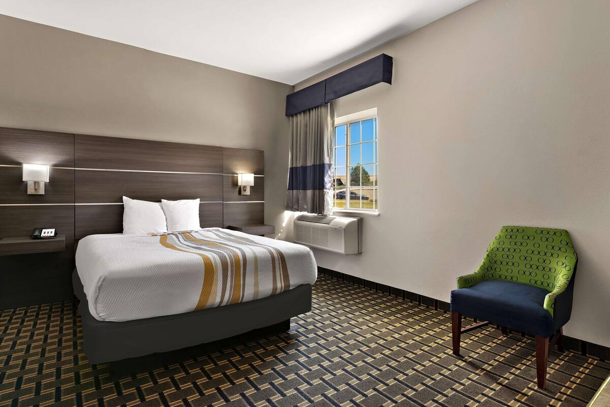 Ein Hotelzimmer mit Bett, Sessel, Nachttisch, Lampe, einem Fenster mit Vorhängen und einem gemusterten Boden.