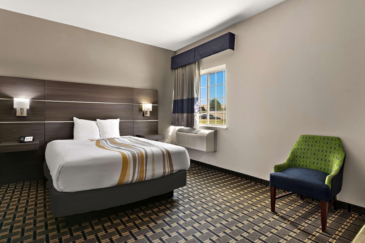 A hotel room with a bed, a chair, a nightstand, a lamp, a window with curtains, and a patterned floor.