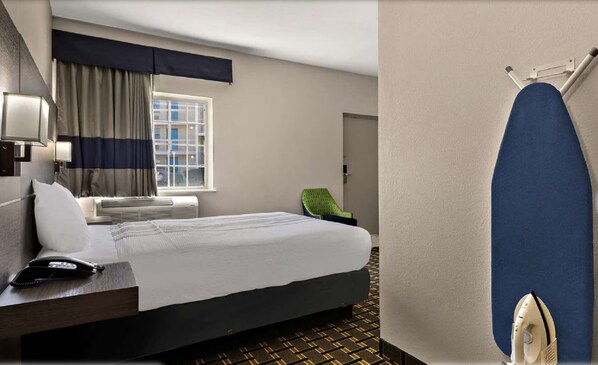 Ein Hotelzimmer mit einem Bett, einem Schreibtisch mit Telefon, einem Sessel, einem Fenster mit Vorhängen und einem wandmontierten Bügelbrett.