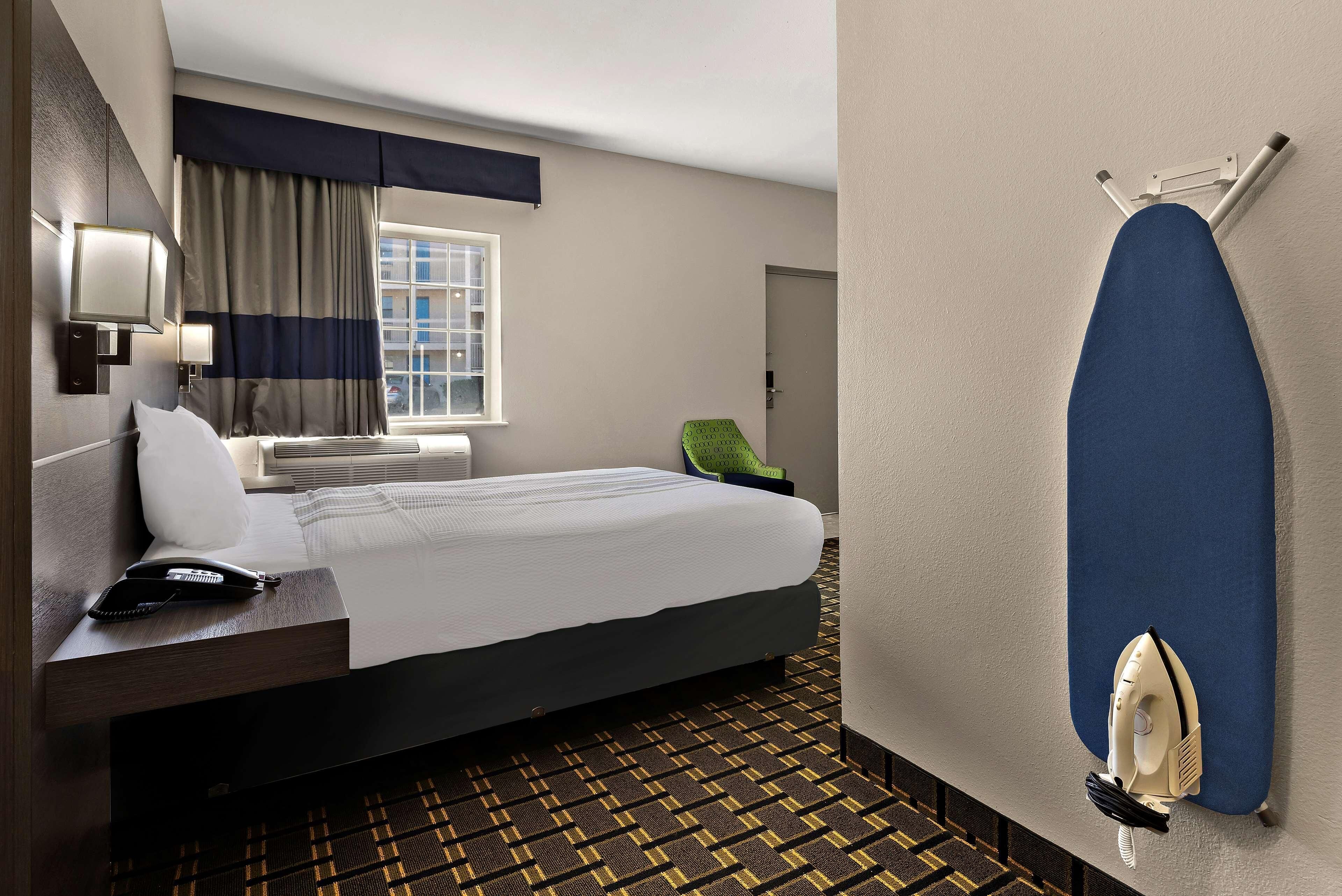 A hotel room with a bed, a desk with a phone, a wall-mounted ironing board, and a patterned carpet.
