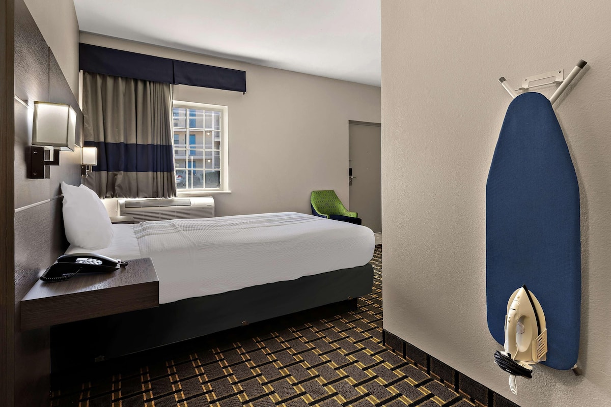 Ein Hotelzimmer mit einem Bett, einem Schreibtisch mit Telefon, einem an der Wand befestigten Bügelbrett und einem gemusterten Teppich.