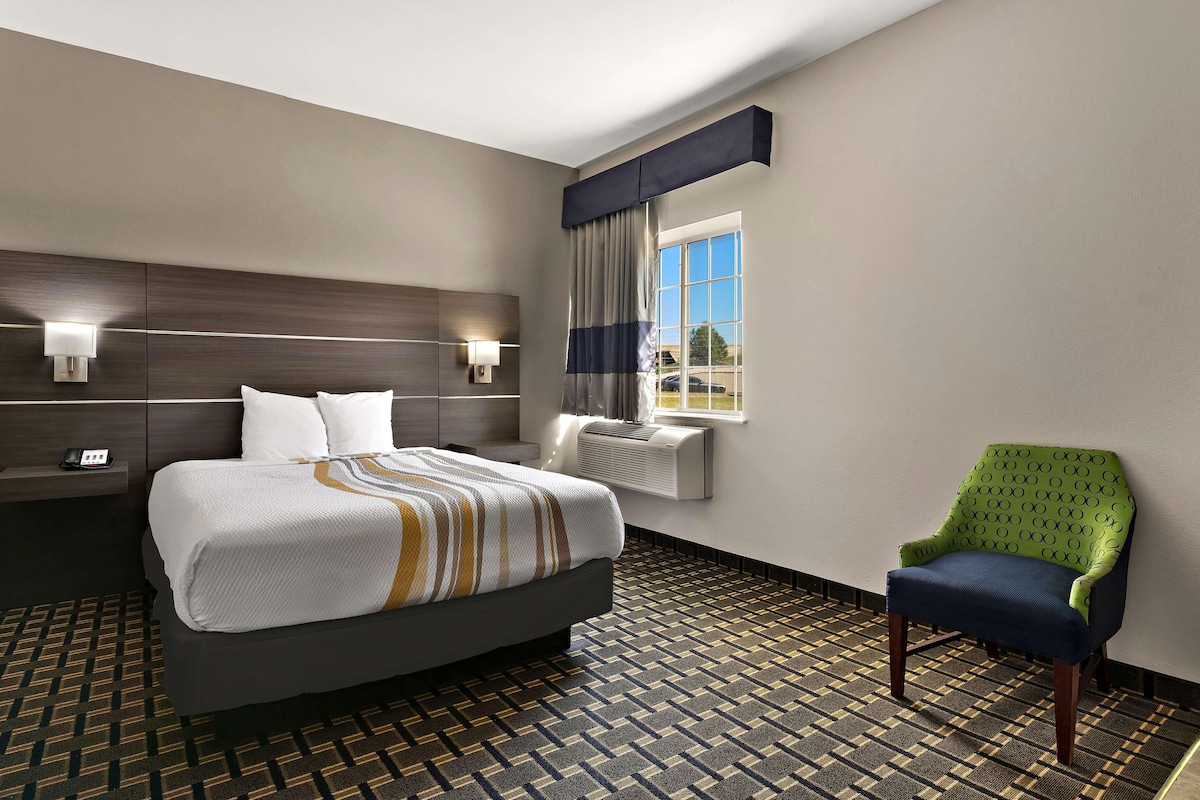 Ein Hotelzimmer mit Bett, Sessel, Nachttisch, Lampe, einem Fenster mit Vorhängen und einem gemusterten Boden.