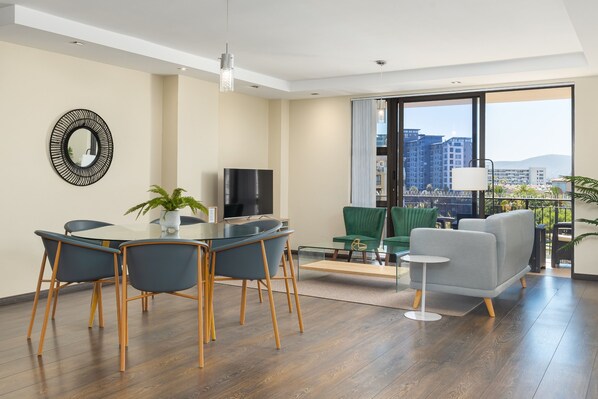 A modern living room with a dining area, a flat-screen TV, and a large window offering a city view.