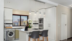 A modern kitchen with a built-in washer and dryer, a large island with bar stools, pendant lighting, and a view through the window.
