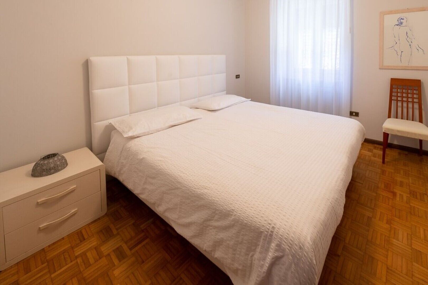 Quarto com cama, mesa de cabeceira, cadeira e um quadro na parede.
