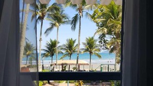 A room with a view of a beach, palm trees, and a thatched-roof structure. - Brisa do Mar Barê Praia Hotel (São Sebastião)