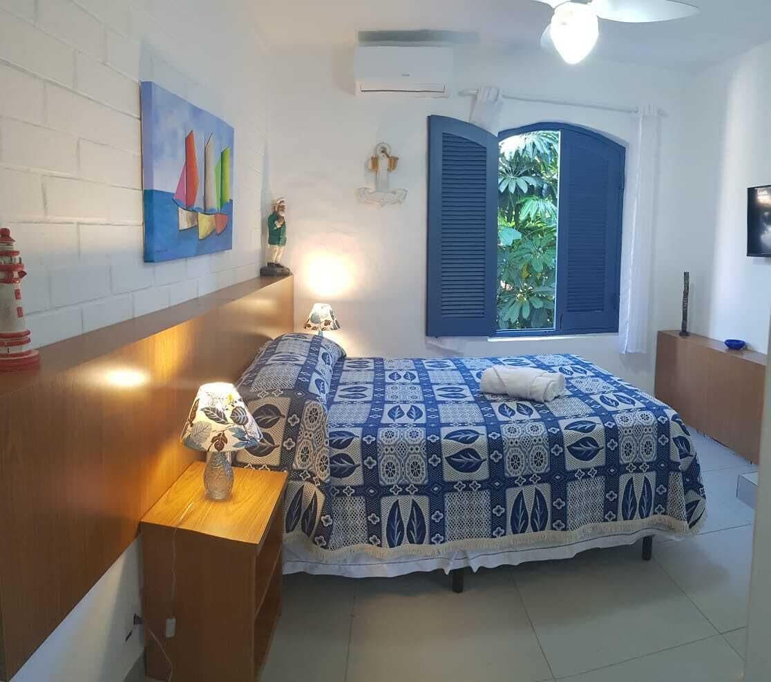 A bedroom with a bed, a nightstand, a lamp, a painting, a window with shutters, and an air conditioning unit.
