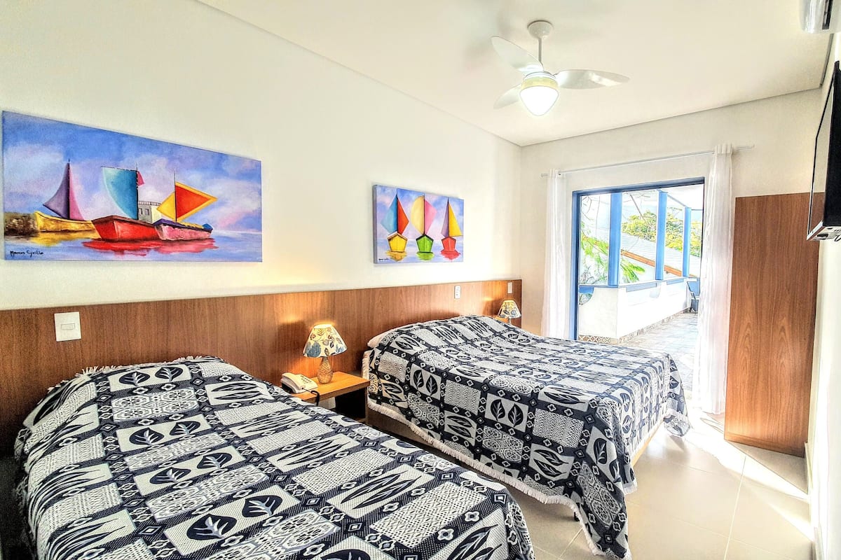 Ein Hotelzimmer mit zwei Betten, einem Deckenventilator, einem Segelboot-Bild, einem Fenster mit Aussicht und einem an der Wand montierten Fernseher.