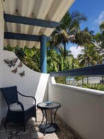 Un balcón con una silla y una mesita, una pared blanca decorada con mariposas, y vistas a palmeras y una playa.