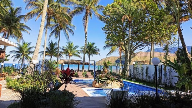 A pool area with palm trees, a rock feature, and a view of the ocean. - Brisa do Mar Barê Praia Hotel (São Sebastião)
