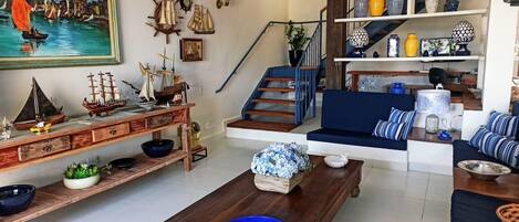 Una sala de estar con una mesa de centro de madera, cuencos azules, un sofá con cojines a rayas, una escalera con barandas azules y un estante con adornos.