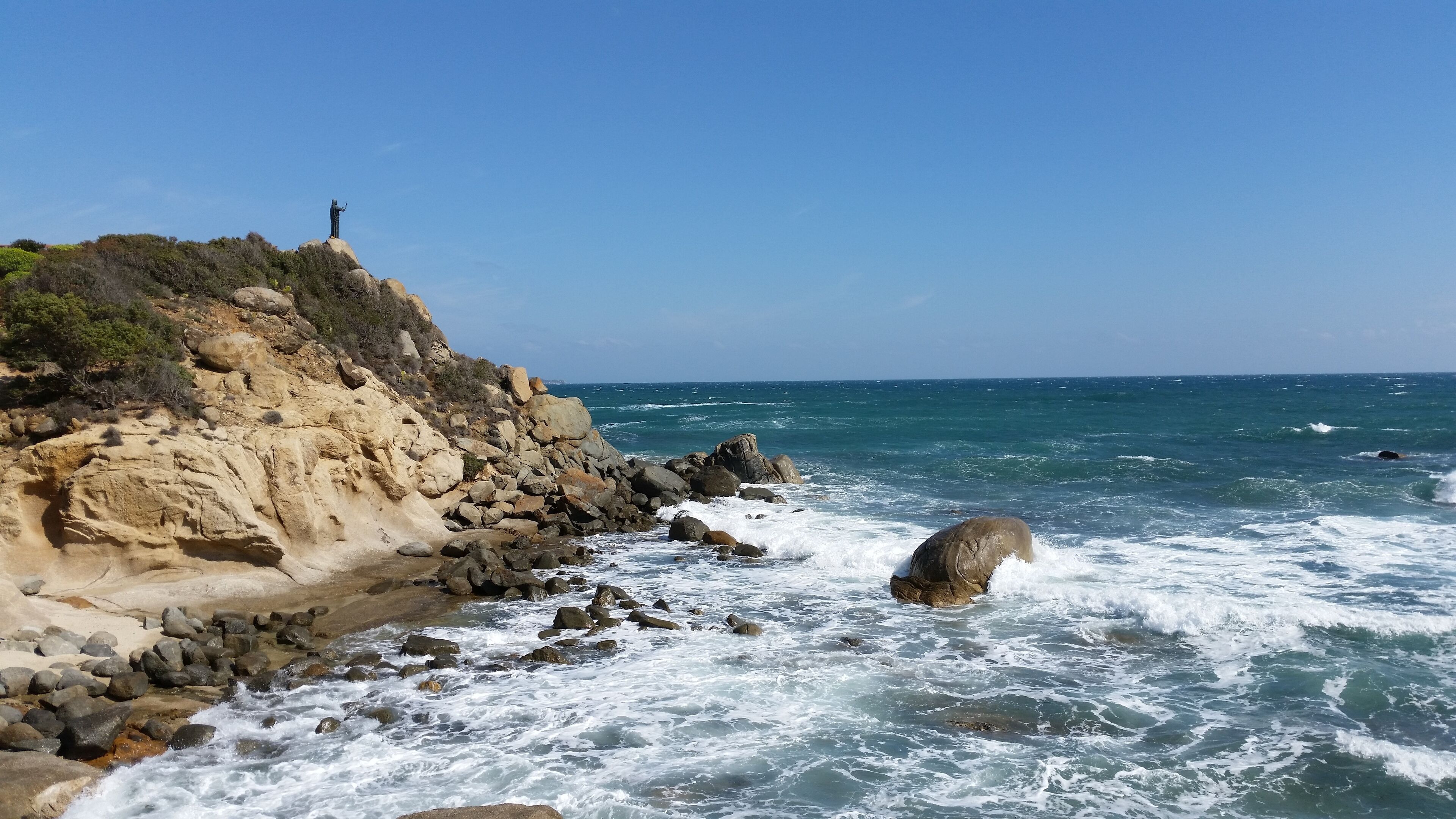 A rocky coastline with a statue on a cliff, waves crashing against the rocks, and a clear blue sky.