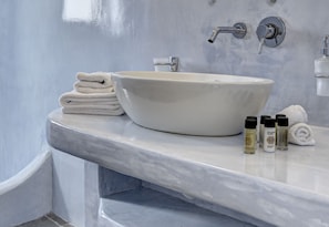 A bathroom with a white sink, two faucets, a towel stack, and three bottles of lotion. - Desiterra Santorini (Santorini)