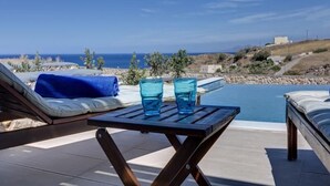 Un espacio al aire libre con mesas iluminado por el sol, vistas al mar, una piscina y cielo despejado.