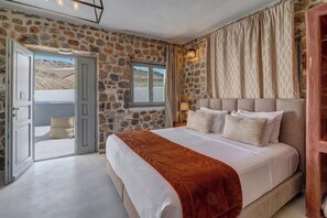 A bedroom with a stone wall, a bed with a brown blanket, a window, and a door leading outside. - Desiterra Santorini (Santorini)