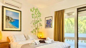 A hotel room with a bed, a large window with curtains, a wall-mounted air conditioner, a framed painting of a beach, and a potted bamboo plant. - Villasonboard Dock Villa 3bed Sleeps8 Porto Hydra (Ermionida)