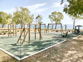 Un parque infantil con columpios y estructuras de escalada, rodeado de árboles y vistas al mar.