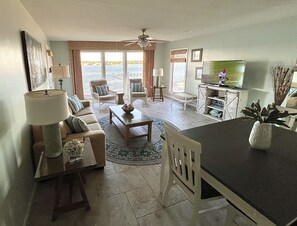 Uma sala de estar ampla com sofá, mesa de centro e cadeiras, área de jantar com mesa e cadeiras, cozinha com televisão e uma janela grande com vista para a água.