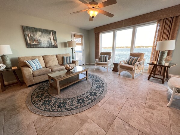 A spacious living room with a large window offering a view of the water, a ceiling fan, and a painting on the wall.