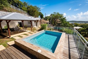 Een zwembad met een houten terras, een overdekte zithoek en uitzicht op een meer en heuvels op de achtergrond.