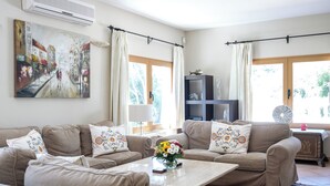 A living room with beige sofas, a coffee table with a vase of flowers, a painting on the wall, and large windows with curtains.