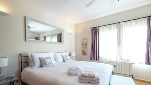 A neatly made bed with white linens and two towels, a bedside table with a lamp, a large mirror, and a window with curtains.