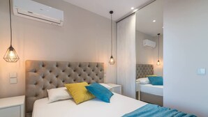 Un dormitorio con cabecera capitonada, cama con ropa de cama blanca y azul, mesita de noche, espejo que refleja la habitación y aire acondicionado de pared.