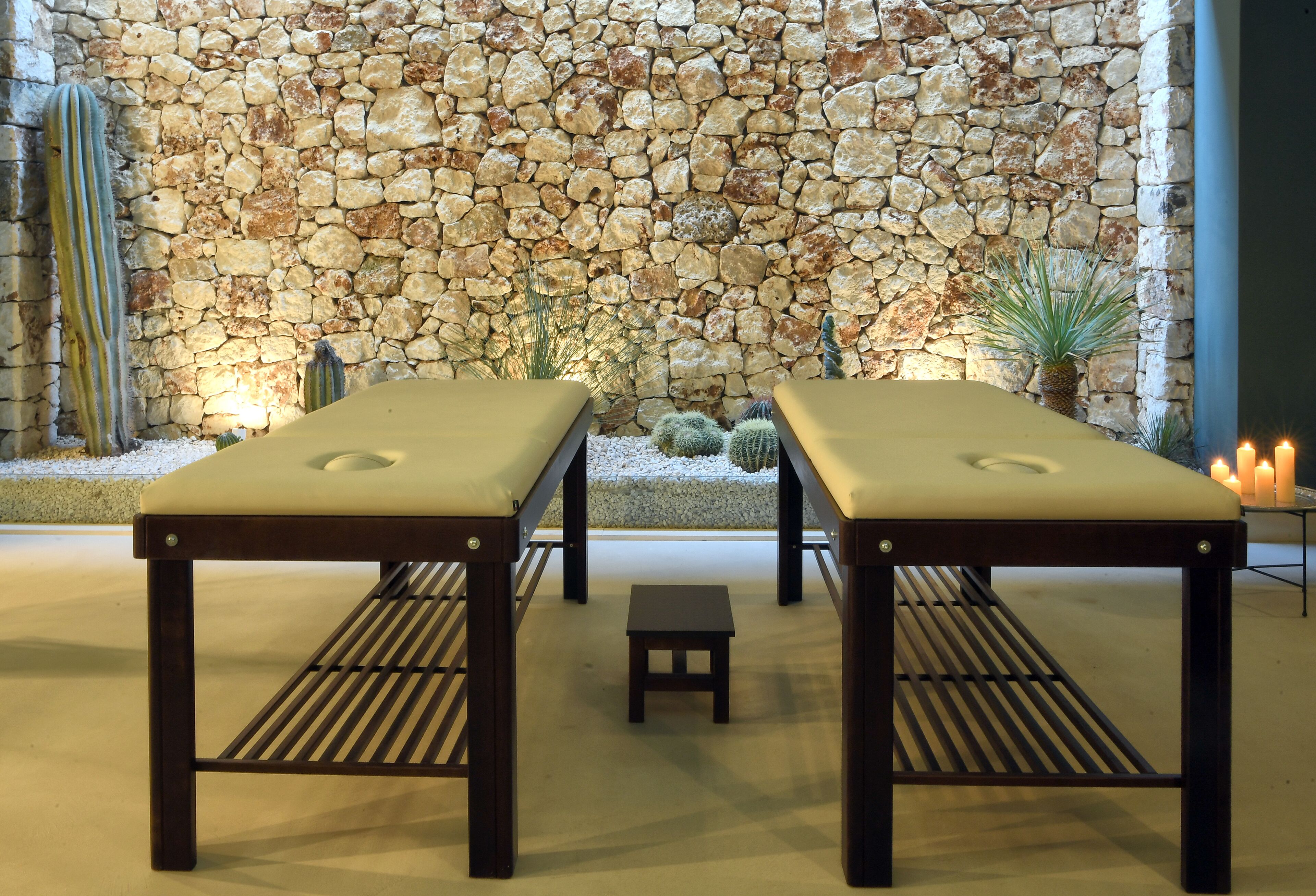 Two wooden massage tables with cushions, a small stool, and a stone wall with cacti in the background.